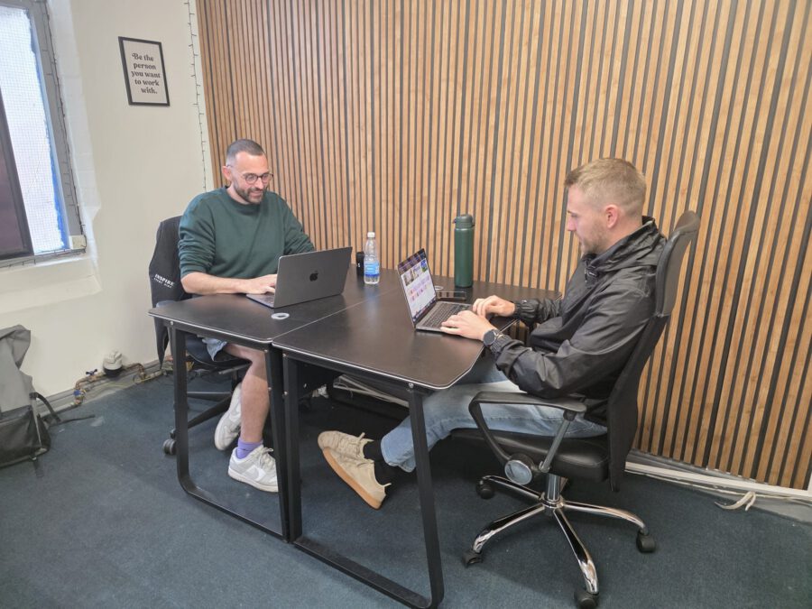 Two members working on laptops at a shared desk in front of the wooden slat wall at Coworking Beeston